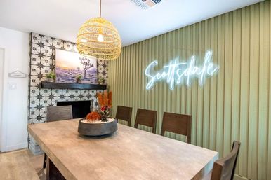 Bright modern Southwestern dining room in Scottsdale with a woven rattan pendant, concrete-style table, wooden chairs, patterned tile fireplace with desert landscape art, and a glowing 'Scottsdale' neon sign on a fluted sage wall.