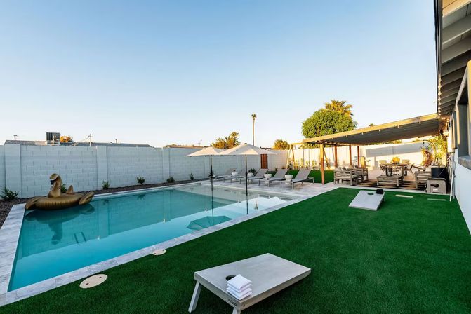 Modern residential backyard with a rectangular pool and gold swan float, sun loungers and umbrellas, pergola-covered outdoor dining area, and artificial turf with cornhole boards