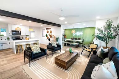 Open-concept living room and kitchen with light hardwood floors, exposed dark beam, white island with bar stools, black lounge chairs, wooden coffee table, green accent dining wall and indoor plants, bright modern farmhouse vibe.