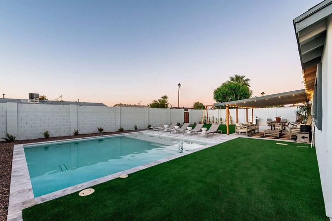 Backyard oasis with rectangular swimming pool, white stone deck and a row of lounge chairs, green turf lawn, pergola-covered patio with string lights and outdoor dining, palm trees and pastel dusk sky