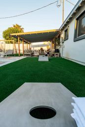 Sunny modern backyard patio with covered pergola, string lights, outdoor seating, potted plants, artificial turf and cornhole boards lined up toward a cozy lounge area