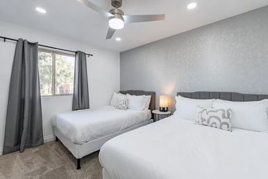 Bright modern guest bedroom with two white-dressed beds, gray textured accent wall, upholstered headboards, ceiling fan, window with gray curtains, bedside table with lamp and carpeted floor