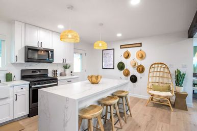 Bright modern coastal-style kitchen with marble waterfall island, three wooden bar stools, woven rattan pendant lights, black stove and microwave, wall-hung hats, rattan accent chair and potted plant.