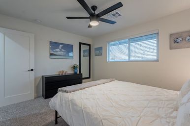 Bright modern bedroom with white comforter on a bed, black ceiling fan, low black dresser with vase and candles, full-length mirror, coastal wall art and a window with horizontal blinds over gray carpet.