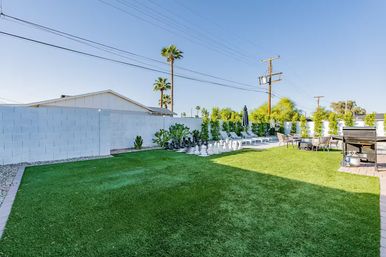 Sunny southwestern backyard with green artificial turf, white block privacy wall, tall palm trees, lounge chairs, oversized outdoor chess set, patio dining area and BBQ grill.