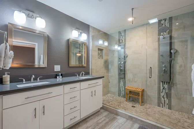 Spa-like contemporary master bathroom with double sink vanity and gray countertop, backlit mirrors, frameless glass walk-in shower with rainfall panel and wooden bench, neutral wall tiles and wood-look floor