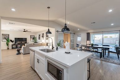 Bright, modern open-concept kitchen and living area with white island and built-in microwave, black pendant lights, farmhouse dining table, sliding glass doors to backyard, and light wood floors