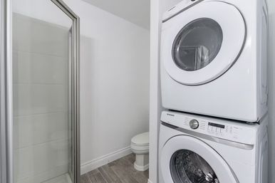 Compact bathroom with space-saving stacked white washer and dryer next to a glass-enclosed shower and toilet on gray wood-look tile floor.