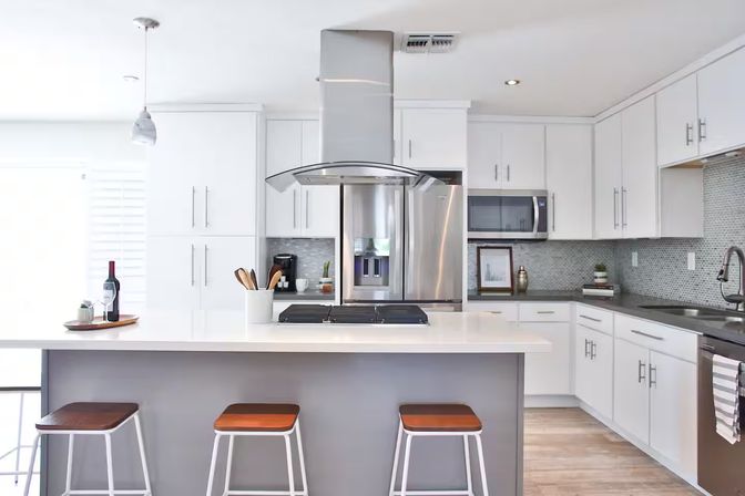 Bright contemporary white kitchen with central island and cooktop, stainless steel range hood and fridge, mosaic tile backsplash and three wooden-top bar stools