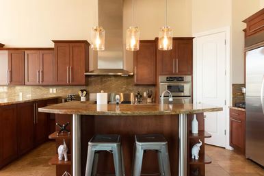 Warm modern kitchen with dark wood cabinets, granite island breakfast bar, two metal stools, stainless steel appliances and hood, glowing glass pendant lights, tiled floor and open shelving accents.