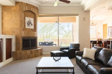 Bright modern living room with tiled corner fireplace, black leather sofas and coffee table, large window overlooking a sunny patio and desert trees