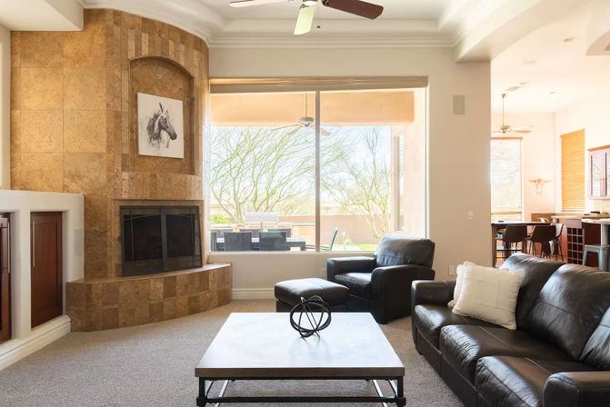 Bright modern living room with tiled corner fireplace, black leather sofas and coffee table, large window overlooking a sunny patio and desert trees