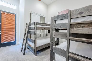 Cozy vacation-rental bunk room with gray wooden twin-over-twin bunk beds, white linens, ladder, carpeted floor, sliding closet and a patio door with wooden blinds.