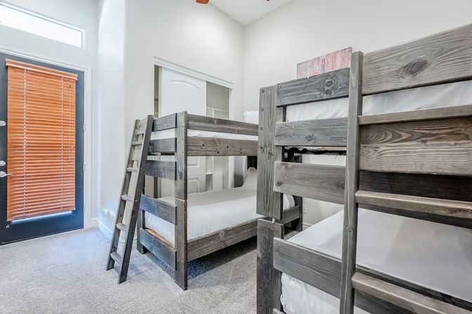 Cozy vacation-rental bunk room with gray wooden twin-over-twin bunk beds, white linens, ladder, carpeted floor, sliding closet and a patio door with wooden blinds.