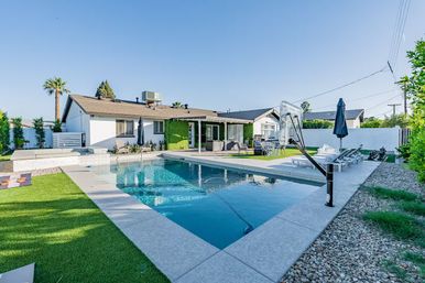 Sunny suburban backyard with a rectangular swimming pool and attached spa, poolside basketball hoop, lounge chairs and umbrellas, outdoor dining and seating on a concrete patio beside a single-story ranch house with artificial turf and palm trees