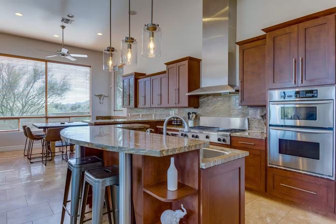 Sunlit contemporary kitchen with a curved granite island and breakfast bar, warm wood cabinets, stainless steel range and double oven, glass pendant lights, metal bar stools, and large windows framing trees.