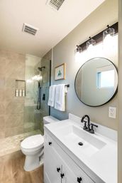 Bright modern home bathroom with white vanity and black faucet, round mirror, glass walk-in shower with rain head, hex tile shower floor and wood-look flooring.