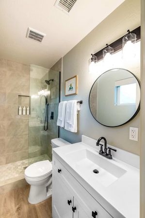 Bright modern home bathroom with white vanity and black faucet, round mirror, glass walk-in shower with rain head, hex tile shower floor and wood-look flooring.