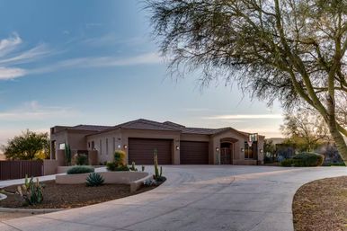 Single-story Southwestern stucco home with three-car garage, curved driveway and desert landscaping with cacti and a large tree at sunset