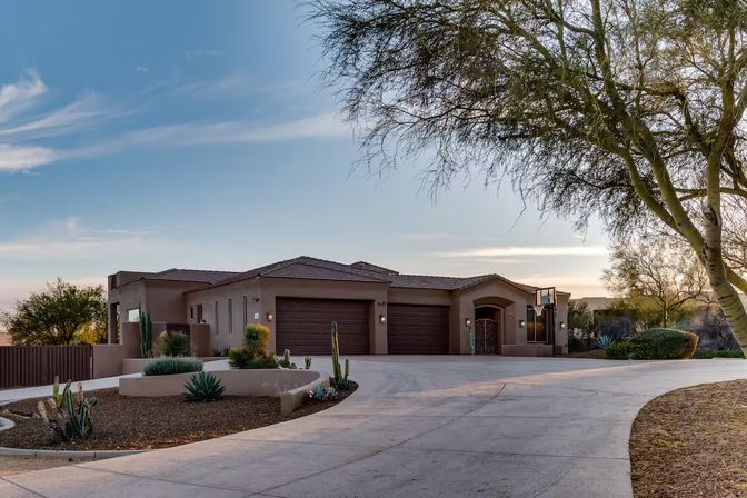 Single-story Southwestern stucco home with three-car garage, curved driveway and desert landscaping with cacti and a large tree at sunset
