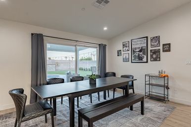 Contemporary dining room in a modern home with a long black table, matching bench and quilted chairs facing sliding glass doors opening to a backyard pool with string lights; gallery wall art, bar cart and gray curtains.
