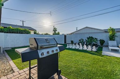 Sunny suburban backyard patio with a stainless steel gas grill on pavers, green lawn featuring an oversized outdoor chess set, white lounge chair, succulents and palm tree against a cinderblock privacy wall.
