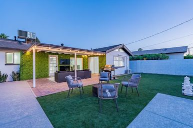 Cozy suburban backyard at dusk with a covered pergola and string lights, outdoor sofa and woven chairs around a coffee table, wall-mounted TV against a green living wall, barbecue grill and manicured lawn enclosed by a white block fence.