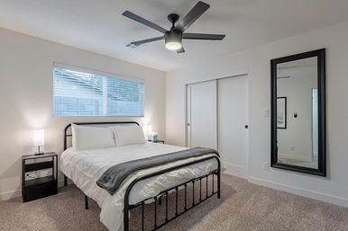 Bright, cozy modern bedroom interior with black metal bed frame, white linens and gray throw, bedside lamps, ceiling fan, sliding closet doors, full-length mirror and carpeted floor