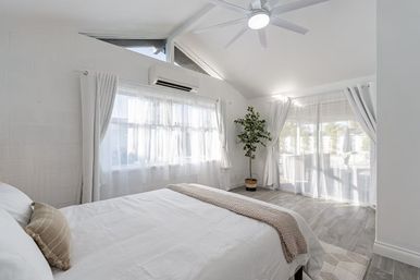 Sunlit modern bedroom with vaulted ceiling and ceiling fan, white linens, sheer curtains, sliding glass doors, potted plant and gray wood-look floors