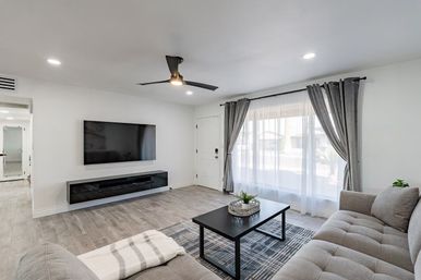 Bright modern living room with wall-mounted flat-screen TV, gray tufted sectional, black coffee table on patterned rug, sheer curtains with gray drapes, wood-look tile floor and a sleek ceiling fan