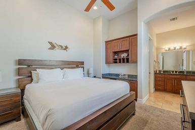 Cozy bright master bedroom with king wooden platform bed and crisp white linens, ceiling fan, built-in wood cabinetry with granite counter and open doorway to an ensuite bathroom with dual-sink vanity.