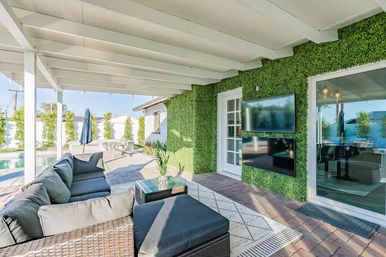 Sunlit modern covered backyard patio with wicker sectional sofa, glass-top coffee table and potted plant, green living wall with two mounted flat-screen TVs, sliding glass door and pool view