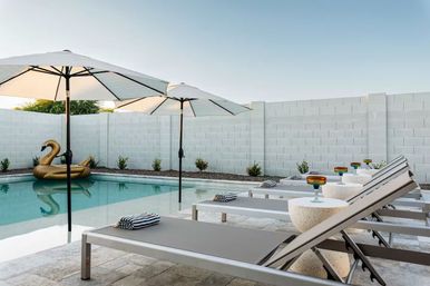 Modern residential backyard pool with a row of gray lounge chairs under white umbrellas, striped towels and colorful cocktail glasses on side tables, and a gold inflatable swan floating in the water.