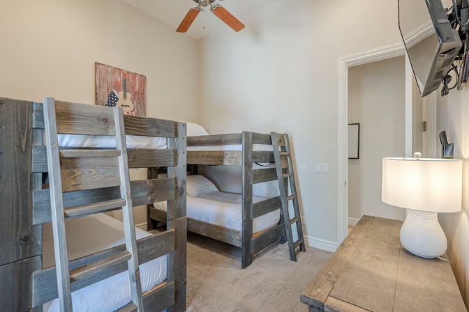 Cozy family-friendly guest room with rustic gray wooden bunk beds, neutral walls and carpet, ceiling fan, wall-mounted TV, wooden dresser with lamp and guitar wall art