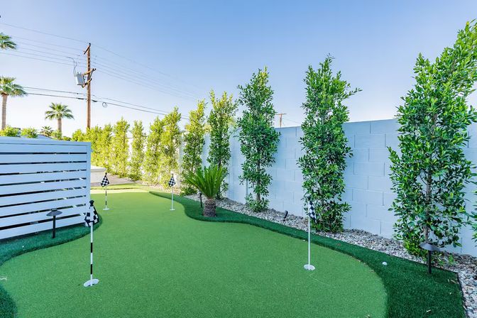 Residential backyard putting green with artificial turf and checkered golf flags, bordered by tall columnar hedges, a white block wall, slatted fence and palm trees under a clear blue sky.