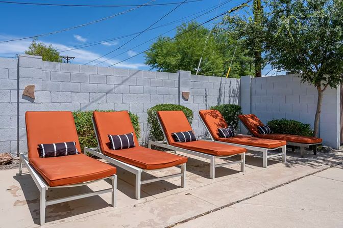 Five orange-cushioned poolside lounge chairs with black-and-white striped pillows lined up against a gray cinderblock wall in a sunny backyard with blue sky and desert landscaping.