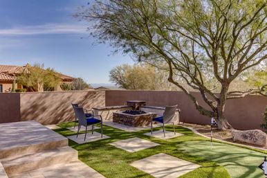 Sunlit Southwestern backyard patio with a mature desert tree, two chairs around a stone fire pit, tiled steps, square stepping stones on turf and a small putting green beside a stucco privacy wall.