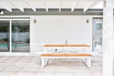 Sunlit minimalist outdoor patio under a white pergola featuring a wooden bench-style dining table, white metal chairs, a small potted succulent centerpiece, and a glass sliding door with a backyard pool view.