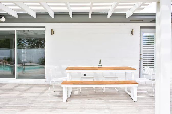 Sunlit minimalist outdoor patio under a white pergola featuring a wooden bench-style dining table, white metal chairs, a small potted succulent centerpiece, and a glass sliding door with a backyard pool view.