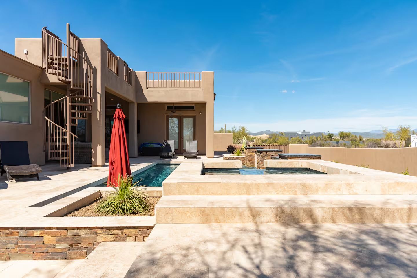 Sunny desert patio of a modern stucco home with stone-tile deck, narrow lap pool and raised spa, spiral metal staircase, red patio umbrella, loungers, and distant mountain views under a clear blue sky