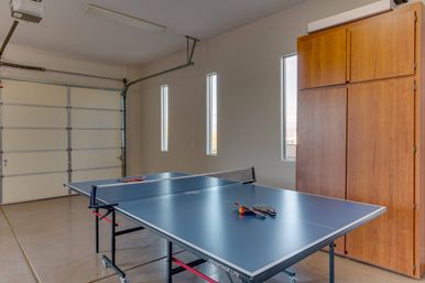 Blue table tennis table set up in a bright residential garage with closed white garage door, three tall narrow windows, wooden storage cabinets, and paddles and an orange ball on the table ready for a match.