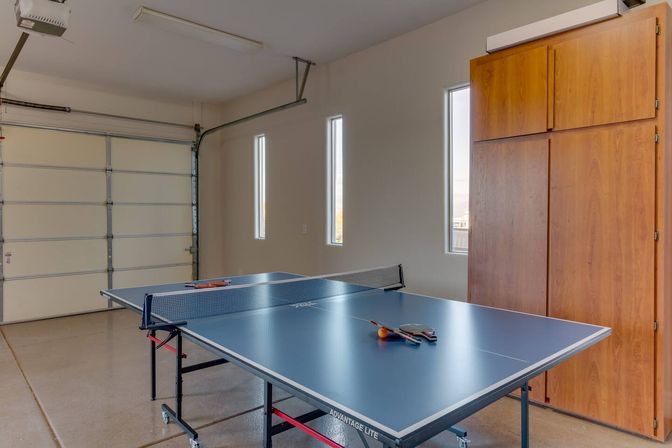 Blue table tennis table set up in a bright residential garage with closed white garage door, three tall narrow windows, wooden storage cabinets, and paddles and an orange ball on the table ready for a match.