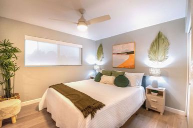 Airy modern bedroom with white quilted bed, green accent pillows and throw, twin bedside tables with lamps, oversized palm-leaf wall decor, potted plant and ceiling fan—bright coastal-inspired retreat.