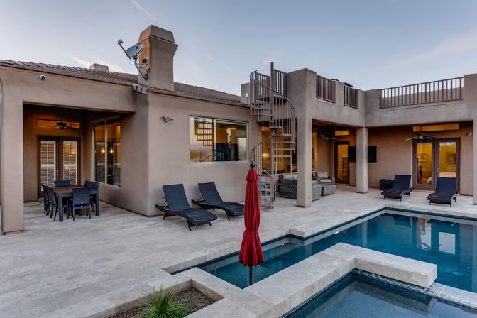 Modern stucco backyard patio with tiled deck, rectangular pool and built-in spa, red closed umbrella, black lounge chairs, outdoor dining set and metal spiral staircase up to a rooftop deck at dusk