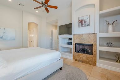 Bright modern master bedroom with white bedding, wooden ceiling fan, tiled floor, built-in shelves and stone-faced fireplace in a neutral, minimalist design