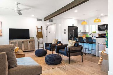 Bright open-plan living room and kitchen with light wood floors, white cabinets, black appliances, breakfast bar with three stools, two black armchairs, navy knit poufs, rattan TV console and pendant lights.