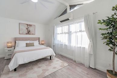 Airy sunlit bedroom with tufted upholstered headboard, crisp white bedding, matching nightstands and warm woven lamps, sheer curtains over large windows, split AC, potted ficus, and geometric rug on light wood floors.