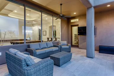 Cozy covered residential patio with gray wicker sectional and ottoman, light-gray cushions, ceiling fan and wall-mounted TV, tiled floor and large glass windows revealing an indoor dining area with a pool table.