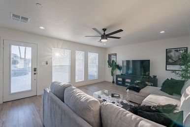 Sunlit modern living room with a gray sectional sofa, glass coffee table, flat-screen TV on a media console, ceiling fan, three large blinds-covered windows, wood-look floors and potted plants.