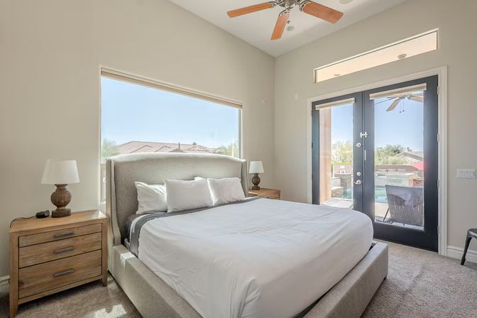 Sunlit modern master bedroom with upholstered platform bed, wooden nightstands and lamps, ceiling fan, large window and glass double doors opening to a patio and pool view